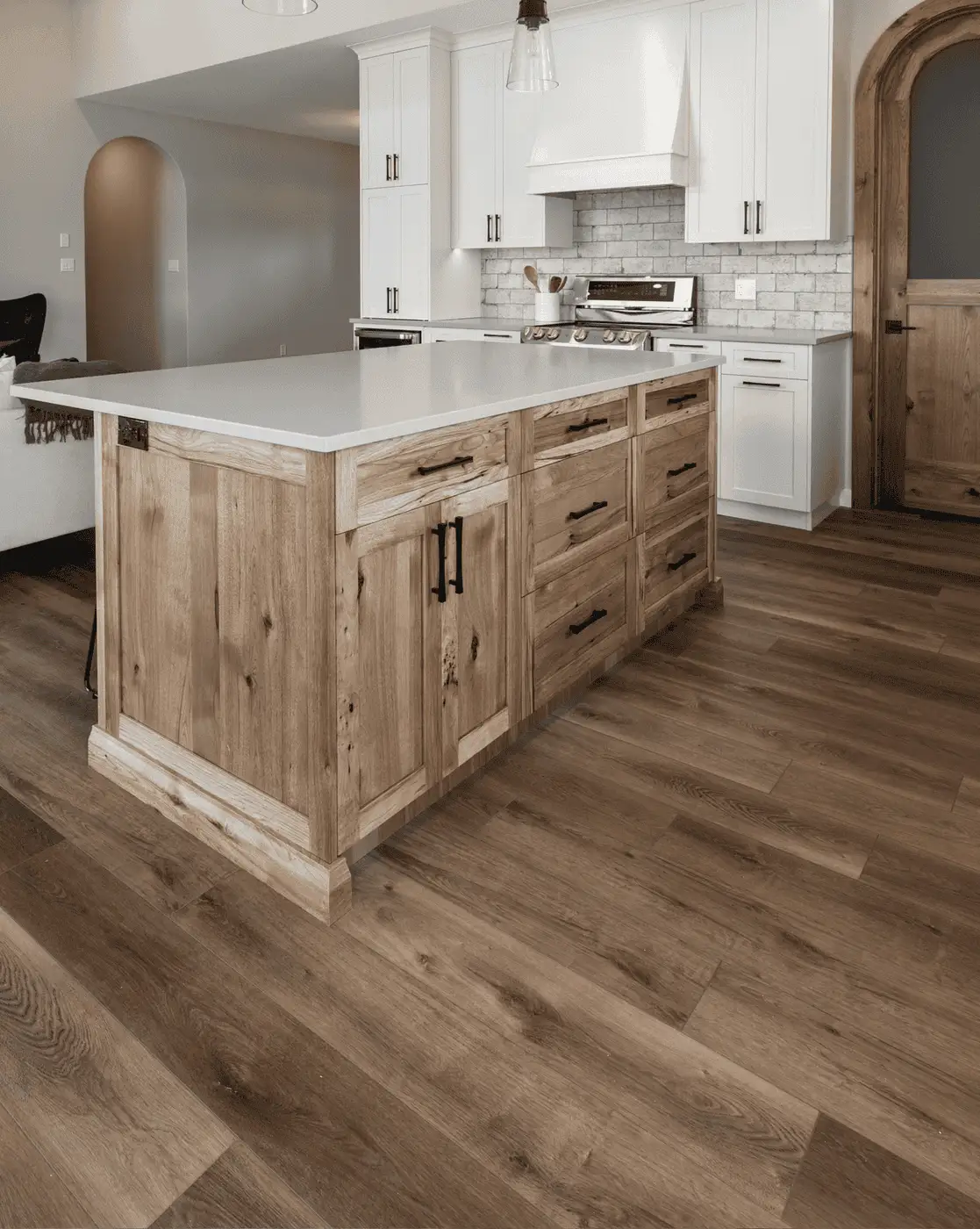 RTM home kitchen in Alberta or Saskatchewan with white cabinets, a large quartz island with natural wood base and drawers, subway tile backsplash, arched wood door, and wide plank floors.