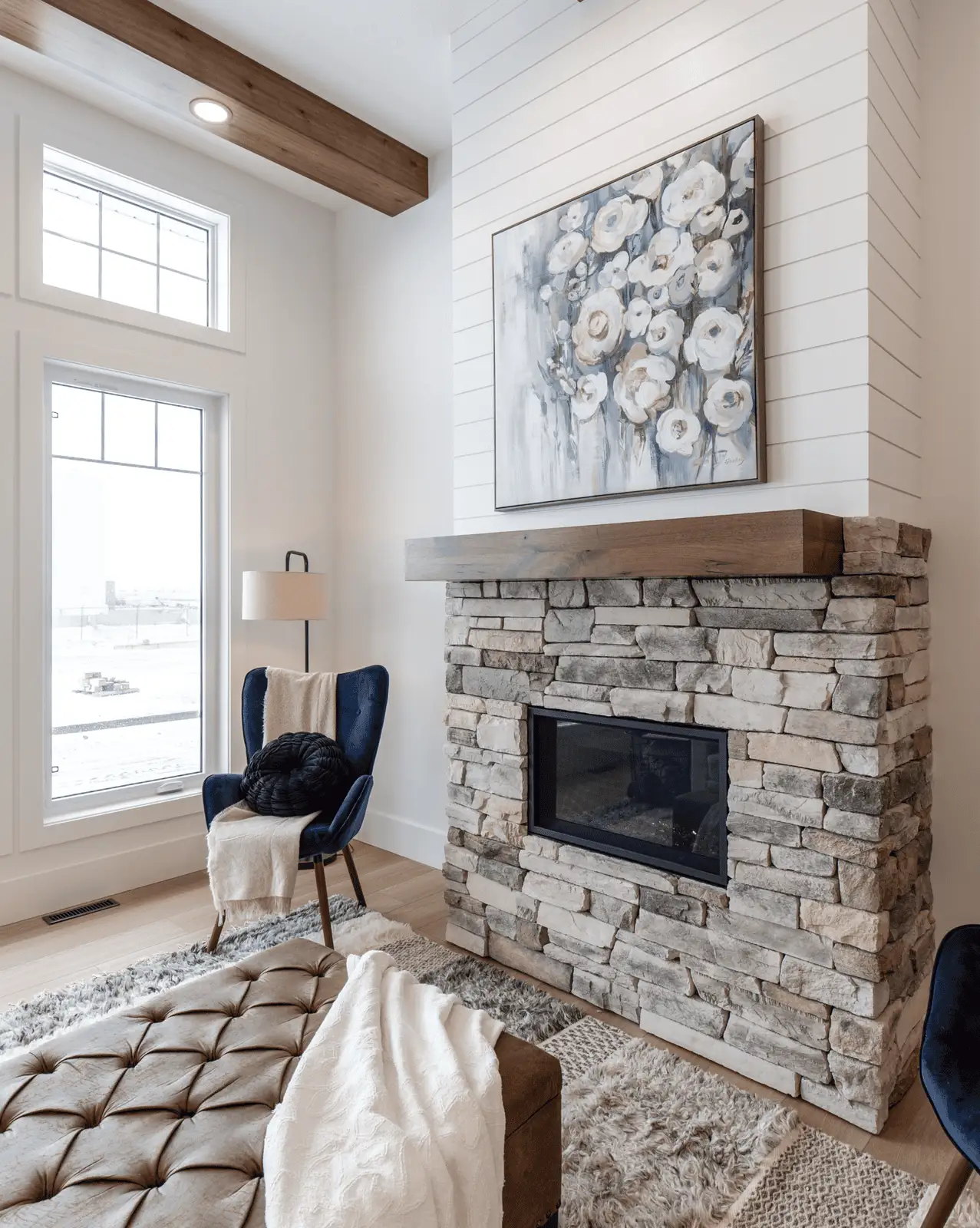 RTM home living room in Alberta or Saskatchewan with a stone fireplace and shiplap wall, wood mantel with floral artwork, large grid window, blue accent chair, and tufted ottoman on layered rugs.