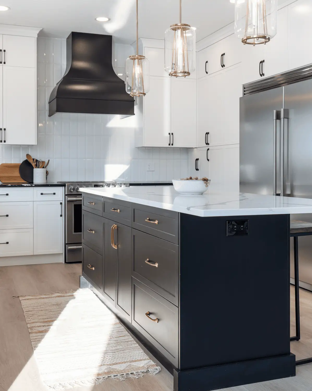 RTM home kitchen in Alberta or Saskatchewan with white cabinets, black feature range hood, quartz counters, dark island with brass pulls, glass pendants, and stainless fridge.