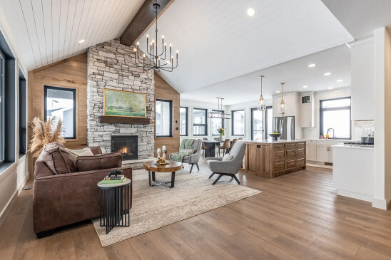 Open-concept RTM home interior featuring a vaulted shiplap ceiling with wood beams, floor-to-ceiling stone fireplace, wide-plank flooring, large black-trim windows, and a custom wood kitchen island with pendant lighting.