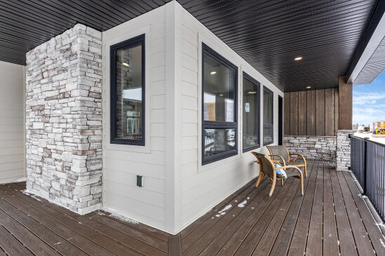 Covered porch of a ready-to-move (RTM) home featuring stone pillars, wood siding, dark trim windows, and composite decking with seating area.