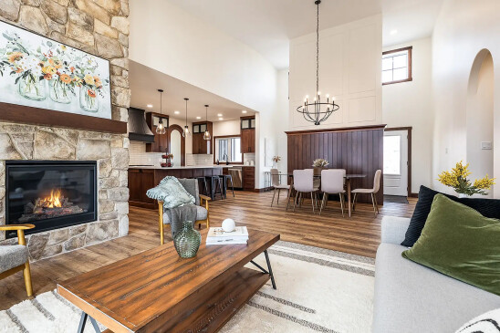 Open-concept living room with stone fireplace, wood coffee table, and modern kitchen and dining area featuring wood cabinetry and pendant lighting.