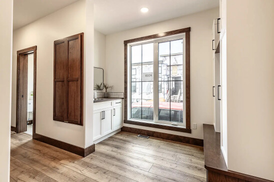 Bright hallway with large window, wood trim, and light wood flooring, featuring white built-in cabinetry with dark hardware and a small vanity area with mirror and plants.