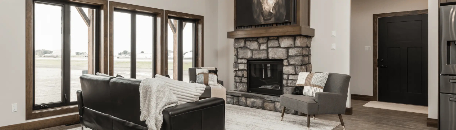 RTM home living room in Alberta or Saskatchewan with large wood-trimmed windows, stone fireplace with horse artwork, black leather sofa, and gray accent chairs.