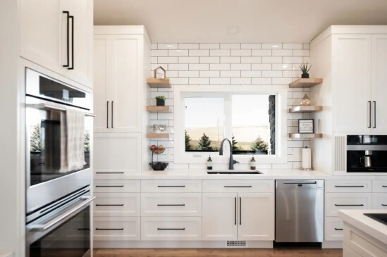 Bright custom kitchen with white shaker cabinets, subway tile backsplash, floating shelves, and stainless steel appliances.