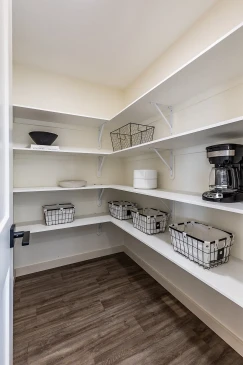 Spacious walk-in pantry in a Saskatchewan home featuring white open shelving with metal baskets, kitchen essentials, and wood-look flooring for a clean and organized design.