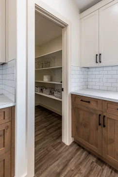 Walk-in pantry in a Saskatchewan home featuring open white shelving with wire baskets, adjacent to a modern kitchen with wood and white cabinetry, subway tile backsplash, and quartz countertops.