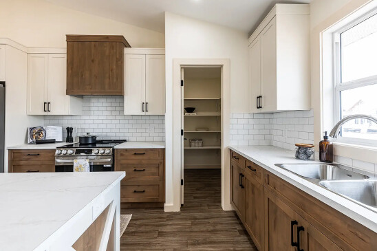 Modern kitchen with two-tone cabinetry, white subway tile backsplash, stainless steel appliances, and a walk-in pantry visible through an open door, designed with warm wood and bright natural light.