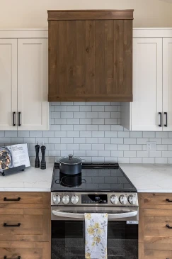 Close-up of a modern kitchen stove with a black cooktop, white subway tile backsplash, wood cabinetry, and a matching wood range hood in a Saskatchewan ready-to-move home.