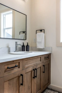 Close-up of a modern bathroom vanity with natural wood cabinetry, white quartz countertop, chrome faucet, black soap dispensers, and a mounted towel beside a large rectangular mirror in a ready-to-move Saskatchewan home.