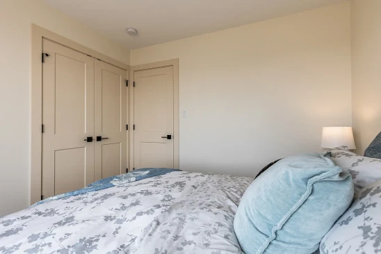 Bright bedroom interior with soft beige walls, matching closet and door, a cozy bed with floral-patterned bedding, blue accent pillows, and a bedside lamp providing warm lighting.