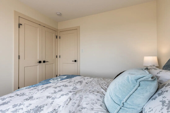 Bright bedroom interior with soft beige walls, matching closet and door, a cozy bed with floral-patterned bedding, blue accent pillows, and a bedside lamp providing warm lighting.