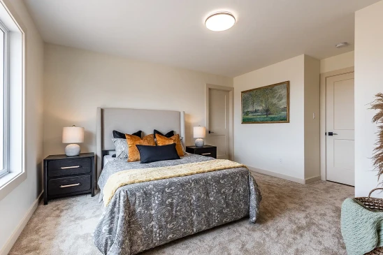 Bright and spacious bedroom in a ready-to-move home featuring a large window, neutral walls, cozy carpeting, a modern upholstered bed with patterned bedding, matching nightstands with lamps, and framed landscape artwork.