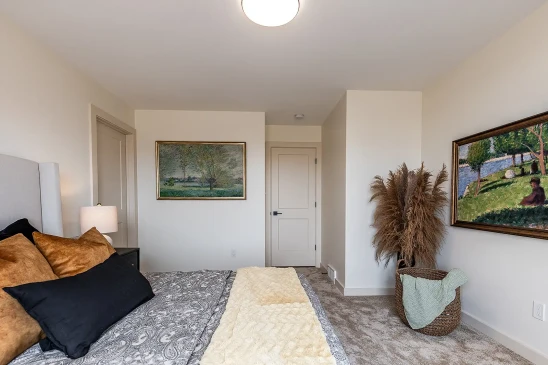 Elegant bedroom in a ready-to-move home featuring neutral walls, framed landscape artwork, a cozy bed with patterned bedding and throw pillows, and a woven basket with decorative pampas grass.
