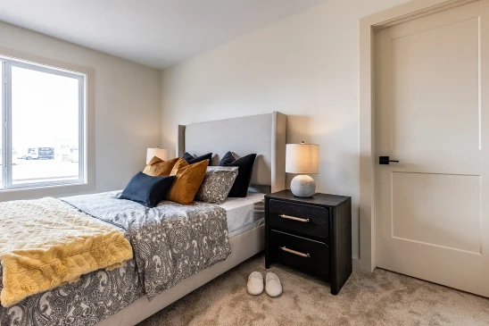 Cozy bedroom in a ready-to-move home featuring a gray upholstered bed with patterned bedding, accent pillows, a dark wood nightstand with a lamp, and a large window letting in natural light.