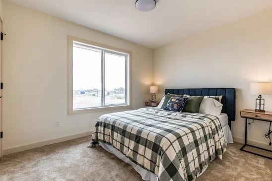 Spacious bedroom in a ready-to-move home featuring a plaid bedspread, cushioned headboard, matching bedside tables with lamps, and a large window providing natural light.