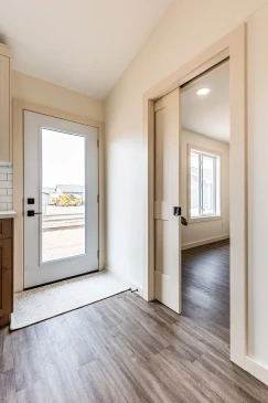 Bright entryway of a ready-to-move home in saskatchewan featuring a glass-paneled exterior door, wood-look flooring, and a sliding interior door leading to a sunlit room.