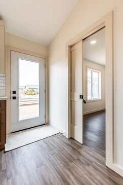 Bright entryway of a ready-to-move home in saskatchewan featuring a glass-paneled exterior door, wood-look flooring, and a sliding interior door leading to a sunlit room.