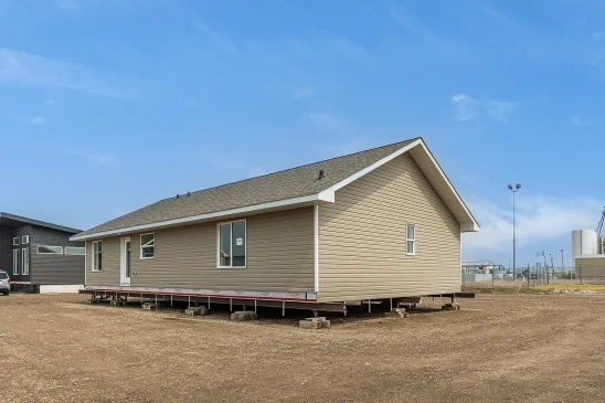 Side view of a tan ready-to-move home in Saskatchewan, elevated on support blocks with white trim, asphalt shingles, and a clear blue sky overhead.