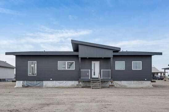 A modern ready-to-move (RTM) home with sleek gray siding and a contemporary flat roof design. The home features a small front porch with white railings and a central front door, accessed by a temporary set of stairs. The exterior includes several large windows that allow natural light to enter the interior. The home is elevated, resting on supports and covered with protective material at the base, indicating it is in the final stages before transport. Surrounding the home are other RTM homes in various stages of construction, and a "Sold" sign is visible next to one of the homes. The sky is partly cloudy, creating a neutral backdrop for the construction site.