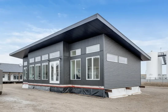 A modern ready-to-move (RTM) home with sleek gray siding and a contemporary flat roof design. The home features a small front porch with white railings and a central front door, accessed by a temporary set of stairs. The exterior includes several large windows that allow natural light to enter the interior. The home is elevated, resting on supports and covered with protective material at the base, indicating it is in the final stages before transport. Surrounding the home are other RTM homes in various stages of construction, and a &quot;Sold&quot; sign is visible next to one of the homes. The sky is partly cloudy, creating a neutral backdrop for the construction site.