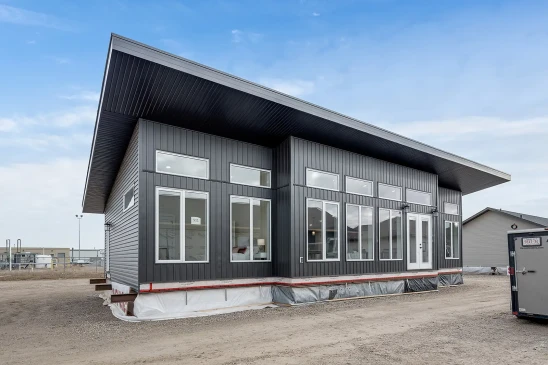 A modern ready-to-move (RTM) home with sleek gray siding and a contemporary flat roof design. The home features a small front porch with white railings and a central front door, accessed by a temporary set of stairs. The exterior includes several large windows that allow natural light to enter the interior. The home is elevated, resting on supports and covered with protective material at the base, indicating it is in the final stages before transport. Surrounding the home are other RTM homes in various stages of construction, and a &quot;Sold&quot; sign is visible next to one of the homes. The sky is partly cloudy, creating a neutral backdrop for the construction site.