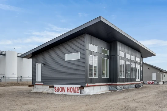 A modern ready-to-move (RTM) home with sleek gray siding and a contemporary flat roof design. The home features a small front porch with white railings and a central front door, accessed by a temporary set of stairs. The exterior includes several large windows that allow natural light to enter the interior. The home is elevated, resting on supports and covered with protective material at the base, indicating it is in the final stages before transport. Surrounding the home are other RTM homes in various stages of construction, and a &quot;Sold&quot; sign is visible next to one of the homes. The sky is partly cloudy, creating a neutral backdrop for the construction site.