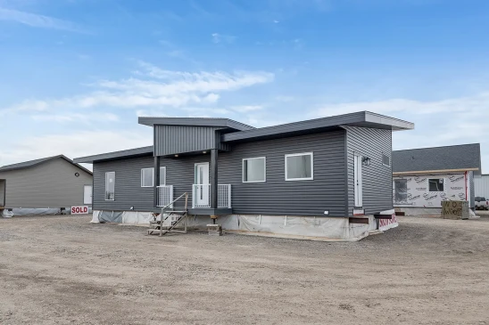 A modern ready-to-move (RTM) home with sleek gray siding and a contemporary flat roof design. The home features a small front porch with white railings and a central front door, accessed by a temporary set of stairs. The exterior includes several large windows that allow natural light to enter the interior. The home is elevated, resting on supports and covered with protective material at the base, indicating it is in the final stages before transport. Surrounding the home are other RTM homes in various stages of construction, and a &quot;Sold&quot; sign is visible next to one of the homes. The sky is partly cloudy, creating a neutral backdrop for the construction site.