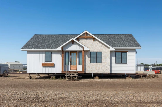 A single-level, ready-to-move (RTM) home with a modern farmhouse exterior. The home features a combination of white siding, stone accents, and wooden trim around the windows and entrance. The front door has a wooden finish with glass panes, and the home sits elevated on a lot, ready for transport to its permanent location. The setting is on a construction lot with clear blue skies overhead.
