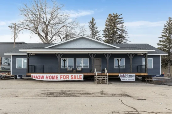 A dark blue ready-to-move (RTM) show home with a large front porch, displayed for sale on a lot. The home features a wide, single-story layout with dark trim, white accents, and a gabled roof. A banner at the front reads &quot;Show Home For Sale,&quot; and stairs lead up to the entrance. The setting is an open area with trees and industrial buildings in the background, showing the home in its transportable stage.