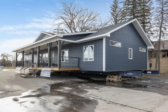 A dark blue ready-to-move (RTM) show home with a large front porch, displayed for sale on a lot. The home features a wide, single-story layout with dark trim, white accents, and a gabled roof. A banner at the front reads &quot;Show Home For Sale,&quot; and stairs lead up to the entrance.