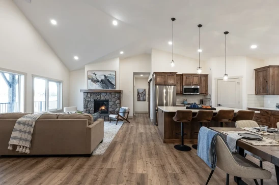 A spacious and modern interior of a ready-to-move (RTM) home featuring an open-concept living room and kitchen. The living area includes a cozy stone fireplace, beige sectional sofa, and large windows that allow natural light to flood the space. The kitchen is equipped with wooden cabinets, stainless steel appliances, an island with bar seating, and pendant lighting. A dining table is set nearby, completing the warm and inviting atmosphere of this stylish home. The hardwood flooring and vaulted ceiling add to the home&#039;s elegant yet comfortable design.