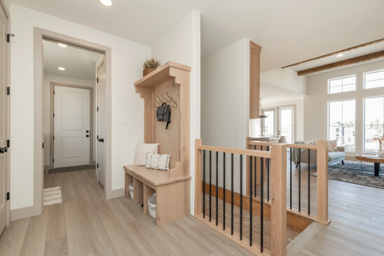 Open and functional foyer with a built-in light wood bench featuring brass hooks and storage baskets, leading to a bright living space with large windows and rustic wood accents, blending practicality and modern design.