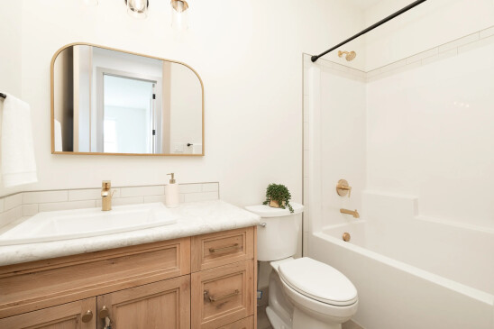Modern bathroom with a light wood vanity, white marble countertop, gold fixtures, a sleek arched mirror, and a clean white bathtub with a black shower curtain rod, creating a bright and elegant design.
